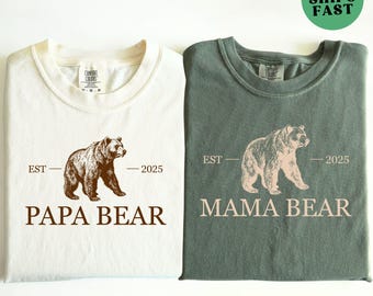 May include: Two t-shirts, one ivory and one moss green, with bear graphics. The ivory shirt says "PAPA BEAR" and the moss green shirt says "MAMA BEAR". Both shirts have "EST - 2025" printed above the bear.
