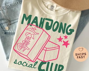 Puede incluir: Camiseta de color marfil con las palabras "MAHJONG social CLUB" en verde y una ilustración rosa de fichas de mahjong y estrellas. La camiseta está doblada sobre unos vaqueros azules, con un collar y un sombrero beige visibles.