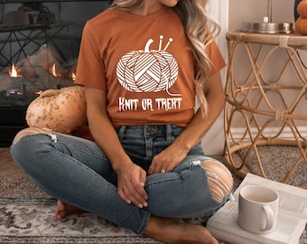 May include: An orange t-shirt with a white graphic of a pumpkin made of yarn and knitting needles. The text "KNIT OR TREAT" is below the pumpkin. The person is wearing blue jeans.