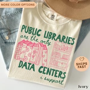 May include: An ivory t-shirt featuring dark green text that reads PUBLIC LIBRARIES are the only DATA CENTERS i support. The design includes pink line art of a row of books and a bookshelf containing books and a globe, printed on the front.