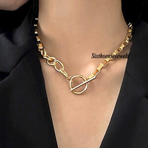 Puede incluir: Collar de cadena dorada con un cierre grande, redondo y de eslabón abierto. El collar lo lleva una persona con una chaqueta negra.