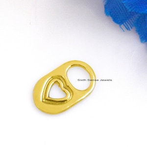 Puede incluir: Un amuleto de color dorado en forma de corazón con un corazón más pequeño cortado en el centro. El amuleto está unido a un anillo y tiene el texto "Sixth Sense Jewels" en el lateral.