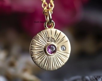 May include: Gold necklace with a round pendant featuring the name "Ziba" engraved on it. The pendant has a sunburst design with a pink gemstone and a small diamond.