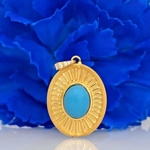 Puede incluir: Un colgante ovalado de oro con una piedra turquesa engastada en el centro. El colgante tiene un diseño decorativo de rayos de sol alrededor de la piedra y una fila de pequeñas piedras transparentes alrededor del borde superior.