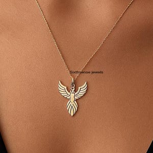 May include: A gold-colored pendant necklace featuring a phoenix design. The pendant is suspended from a delicate chain. The phoenix has outstretched wings and detailed feathering. The necklace is worn against a dark background.