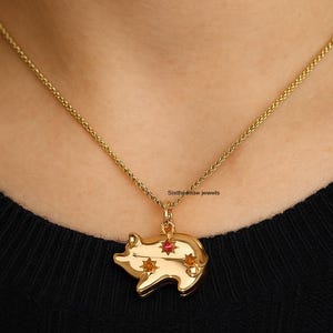 Puede incluir: Un collar dorado con un colgante de cerdo. El colgante tiene una gema roja y dos gemas naranjas en forma de estrella. El collar se lleva sobre una camiseta negra.