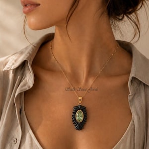 May include: A gold necklace with a black pendant featuring a light green oval stone. The pendant has a decorative black border. The necklace is worn by a person wearing a beige button-down shirt. The text "Sixth Sense Jewels" is visible on the pendant.