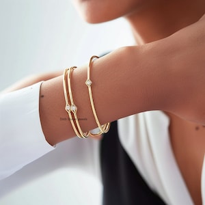 Puede incluir: Tres pulseras de oro con pequeños detalles de diamantes. Las pulseras están apiladas en la muñeca de una persona. El texto "Sixth Sense Jewels" es visible en la pulsera inferior.