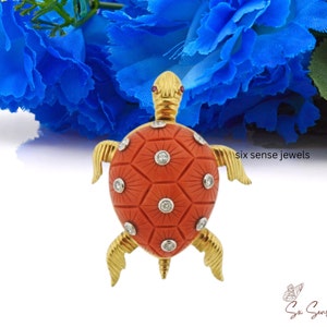 Può includere: Un broche a forma di tartaruga in oro con un guscio di corallo intagliato e accenti di diamanti. La tartaruga è posizionata su uno sfondo floreale blu.