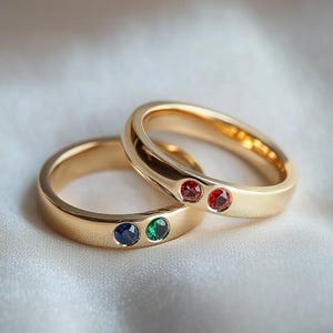 Pode incluir: Duas alianças de ouro com pedras preciosas. A aliança de cima tem duas pedras preciosas vermelhas. A aliança de baixo tem uma pedra preciosa azul e uma pedra preciosa verde.