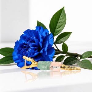 May include: A gold-toned snake-shaped jewelry piece featuring a green stone segment, a pale pink stone segment, and a textured gold tail. The piece is positioned in front of a vibrant blue carnation flower with green leaves and displays the text Sixth Snake Jewels.