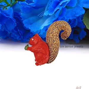 May include: A red squirrel brooch with a gold-colored tail, holding a green nut in its paws. The brooch is made of enamel and metal and has a rhinestone eye. The text "six sense jewels" is visible in the image.