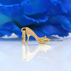 May include: A gold high heel shoe charm with clear sparkling stones. The charm is on a white surface with a blue flower in the background. Sixth Sense Jewels is written on the surface.