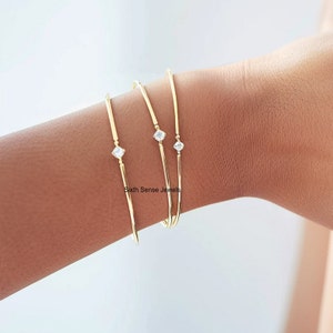 Puede incluir: Tres pulseras de brazalete de oro, cada una con una sola piedra de circonita cúbica transparente. Las pulseras están apiladas en la muñeca de una persona.