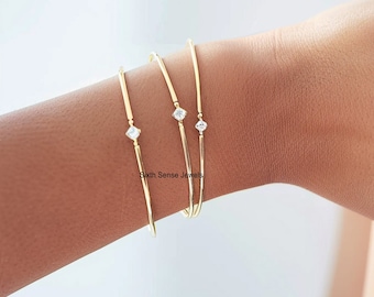 Puede incluir: Tres pulseras de brazalete de oro, cada una con una sola piedra de circonita cúbica transparente. Las pulseras están apiladas en la muñeca de una persona.
