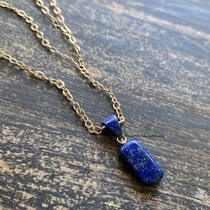 May include: A gold chain necklace with a pendant featuring a dark blue lapis lazuli stone. The stone is rectangular in shape and has a rough, natural texture. The pendant is attached to the chain with a gold bail.