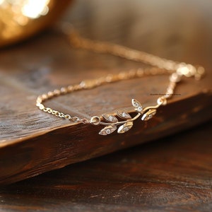 Puede incluir: Un delicado brazalete de cadena dorada con un pequeño diseño de hoja brillante. La cadena está hecha de pequeños círculos unidos y la hoja está adornada con pequeñas piedras transparentes.