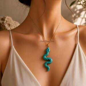 Può includere: Una collana dorata con un ciondolo a forma di serpente turchese. Il ciondolo a forma di serpente, in una vibrante tonalità turchese, è appeso a una sottile catena d'oro. La collana è indossata su un top dai toni neutri.