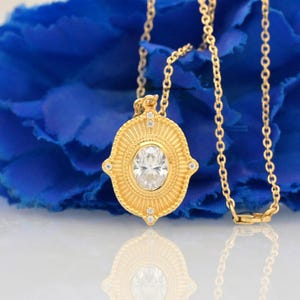 Può includere: Una collana con pendente dorato con una gemma ovale e trasparente al centro. Il pendente presenta un design dettagliato a forma di raggio di sole ed è impreziosito da piccole gemme trasparenti. Anche la catena della collana è dorata.
