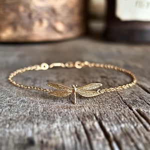 Könnte beinhalten: Ein zartes, goldfarbenes Armband mit einem detaillierten Libellen-Anhänger. Die Libelle hat komplizierte Flügelmuster und ist der Mittelpunkt des Armbands. Die Kette ist fein und das Armband wird auf einer rustikalen Holzoberfläche präsentiert.