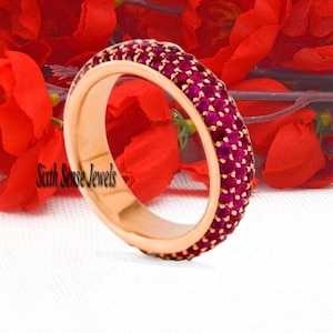 Pode incluir: Um anel de banda de ouro rosa com uma fileira de pequenas pedras preciosas de cor vermelha escura ao redor de toda a banda. O anel está colocado sobre um fundo de tecido vermelho.