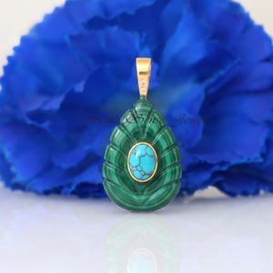 May include: A teardrop-shaped pendant featuring a carved green malachite base with a central oval turquoise cabochon. The piece is set in gold with a gold bail at the top, which includes two small embedded clear stones. The design has ribbed edges.