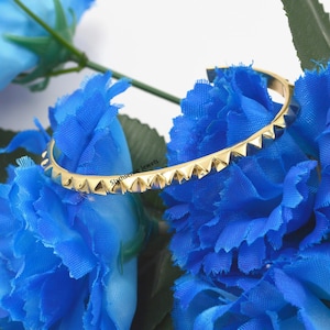 Może przedstawiać: Złota bransoletka z rzędem trójkątnych kolców. Bransoletka leży na niebieskim tle.