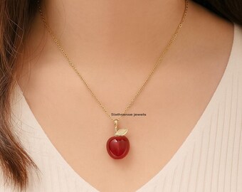 Puede incluir: Un collar dorado con un colgante en forma de manzana roja. La manzana tiene una pequeña hoja dorada adjunta. El collar lo lleva una persona con una camiseta blanca. El texto "Sixthsense jewels" es visible.