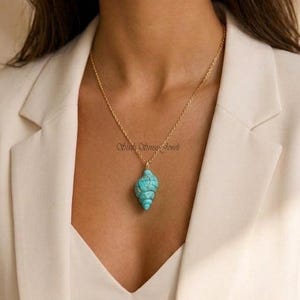 May include: A gold-colored chain necklace with a turquoise seashell pendant. The pendant is a detailed carving of a spiral shell. The necklace is worn over a cream-colored blazer.