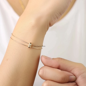 May include: A gold bangle bracelet with a small rectangular charm set with clear cubic zirconia stones. The bracelet is open and has a delicate, minimalist design. "Sixth Sense Jewels" is printed on the bracelet.