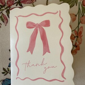 May include: A white card with a pink bow and the words "thank you" in cursive. The card has a scalloped edge and a pink border. The background is a floral pattern.