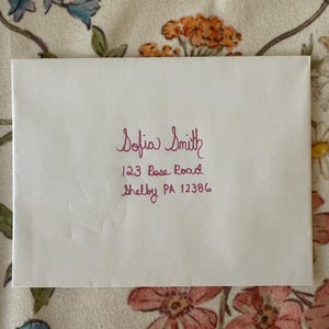May include: A white envelope with handwritten text in a dark pink hue. The text reads "Sofia Smith" followed by a street address and city, "123 Rose Road, Shelby PA 12386". The envelope is on a floral patterned fabric.