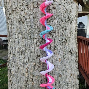May include: A long, colorful crocheted spiral hanging from a tree trunk. The spiral is made of pink, purple, and blue yarn.