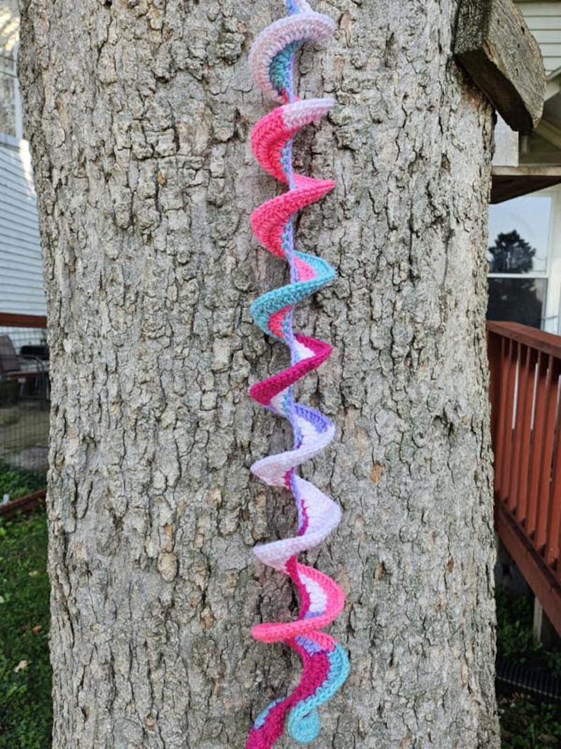 Pu&ograve; includere: Una lunga spirale all'uncinetto colorata appesa a un tronco d'albero. La spirale &egrave; fatta di filato rosa, viola e blu.