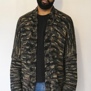 May include: A long, open-front cardigan in a camouflage pattern of green, brown, and black. The knitted sweater is worn over a black t-shirt and blue jeans. The cardigan has a relaxed fit and long sleeves.