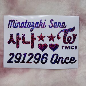 May include: A white sticker with holographic text and designs. The sticker features the name "Minatozaki Sana", Korean characters, stars, hearts, and the word "TWICE". The number "291296" and the word "Once" are also included.