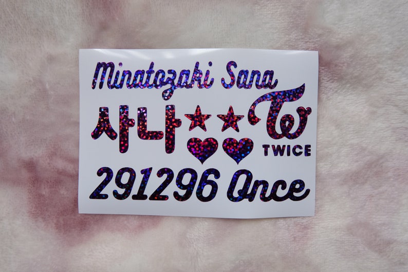 May include: A white sticker with holographic text and designs. The sticker features the name "Minatozaki Sana", Korean characters, stars, hearts, and the word "TWICE". The number "291296" and the word "Once" are also included.