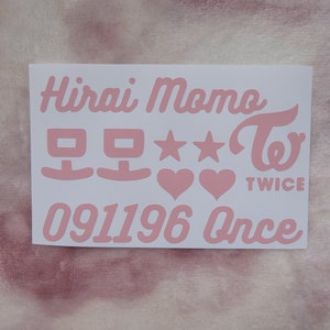 May include: A white sticker with pink text featuring the name "Hirai Momo", Korean characters, stars, hearts, the word "TWICE", the number "091196", and the word "Once". The sticker is on a pink and white background.