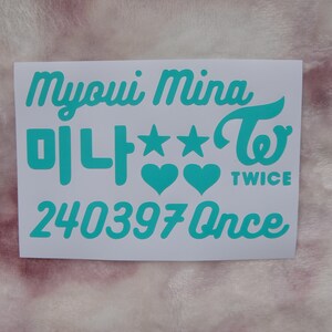 May include: A white sticker with teal text. The text includes the name "Myoui Mina", Korean characters, stars, hearts, the word "TWICE", and the number "240397Once". The sticker is on a pink, textured surface.