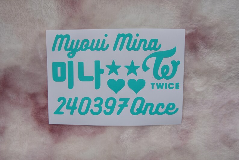 May include: A white sticker with teal text. The text includes the name "Myoui Mina", Korean characters, stars, hearts, the word "TWICE", and the number "240397Once". The sticker is on a pink, textured surface.