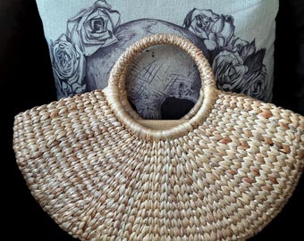 Könnte beinhalten: Eine gewebte Strohhandtasche mit rundem Griff. Die Tasche hat eine fächerartige Form und besteht aus hellbraunem und beigem gewebtem Material. Im Hintergrund befindet sich ein dekoratives Kissen mit einem Totenkopf- und Rosenmuster.