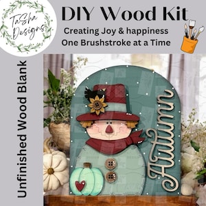 May include: DIY wood kit featuring a wooden scarecrow with a red hat, scarf, and buttons, and a green pumpkin with a heart. The word "Autumn" is displayed in a cursive font. The kit is an unfinished wood blank.