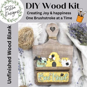 May include: Unfinished wooden cutting board with a heart-shaped cutout at the top. The board has a shelf with a honey jar, a beehive with a bee, and another honey jar. Below the shelf is a sign that says "Bee Kind" in yellow letters with black outlines. The sign has a yellow and black polka dot background.