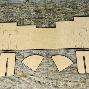 May include: A collection of unfinished, light brown wooden pieces for a craft project. The set includes a long, rectangular base with cut-out slots, two smaller rectangular pieces, and two curved pieces. The pieces are arranged on a wooden surface.