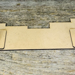 May include: A light brown, rectangular wooden display stand with two vertical slots and a series of cutouts. The stand is designed to hold items and is set against a weathered wood surface.