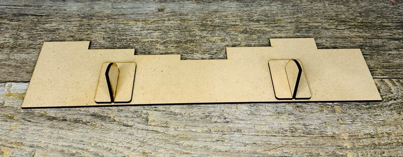 May include: A light brown, rectangular wooden display stand with two vertical slots and a series of cutouts. The stand is designed to hold items and is set against a weathered wood surface.