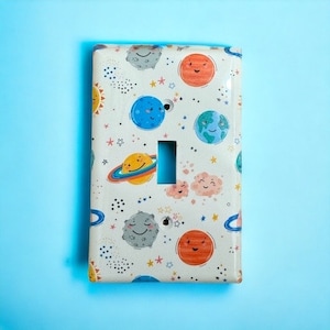 May include: A white light switch cover with a colorful space-themed design. The design features planets, stars, and a sun with smiling faces.