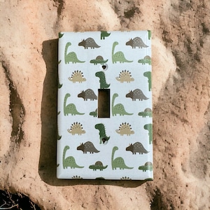 May include: White light switch cover with a pattern of green and brown dinosaurs. The switch cover has a rectangular shape and a white background.