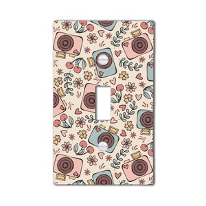 May include: A light switch cover with a vintage camera and floral pattern. The background is a light beige color. The cameras are pink, blue, and brown with a white outline. The flowers are yellow, pink, and blue. There are also small red hearts and stars scattered throughout the pattern.