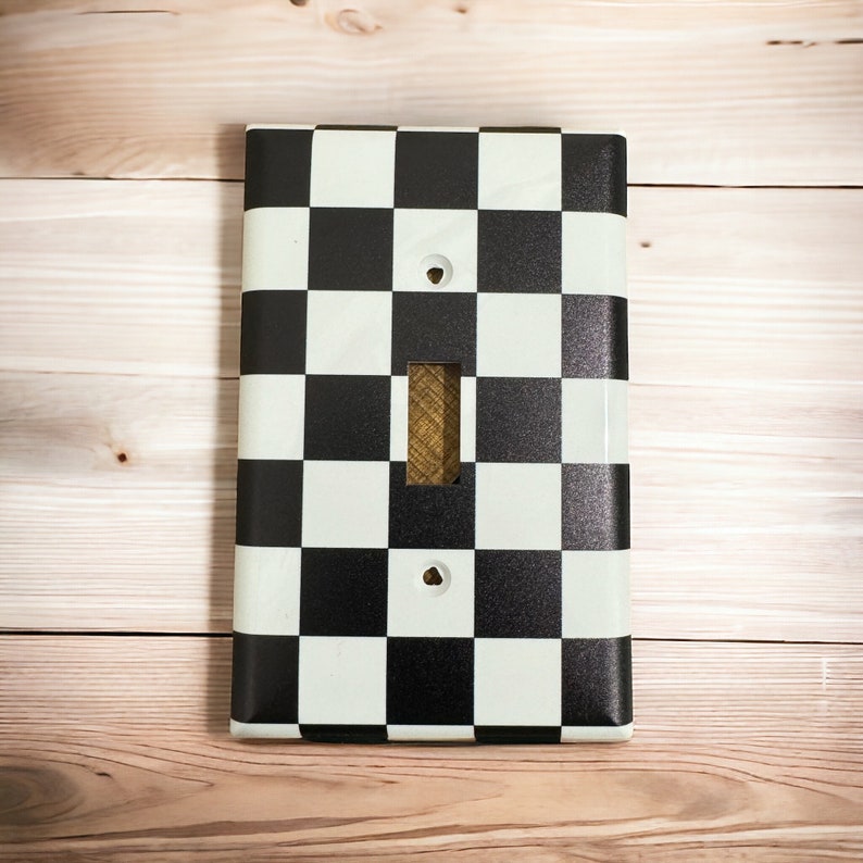 May include: A black and white checkered light switch cover plate. The plate has a standard light switch opening and two small holes for screws.